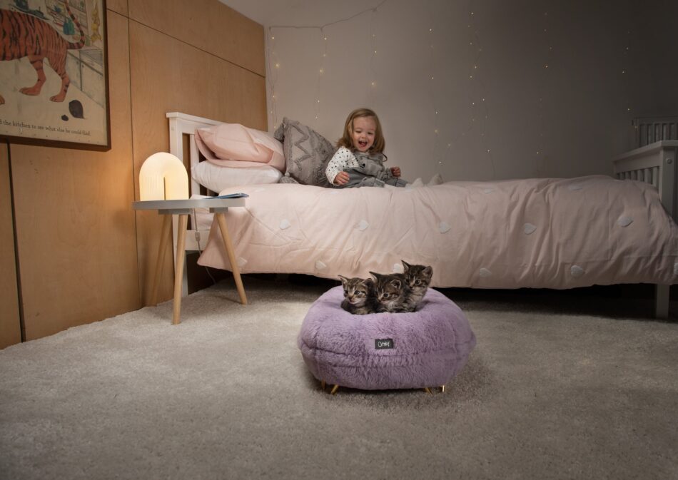 En flicka sitter i en säng och tittar på kattungar på kattbädden Maya Donut i färgen puderlila